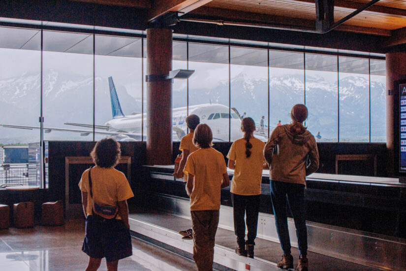 5 people at the airport looking out large windows at a plane and big mountains.