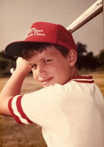 Young boy wearing a white shirt and red hat who is holding a baseball bat above his shoulder, getting ready to swing.