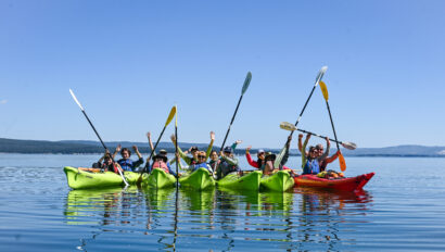 Group of students in kayaks on a blue lake holding their paddles in the air.