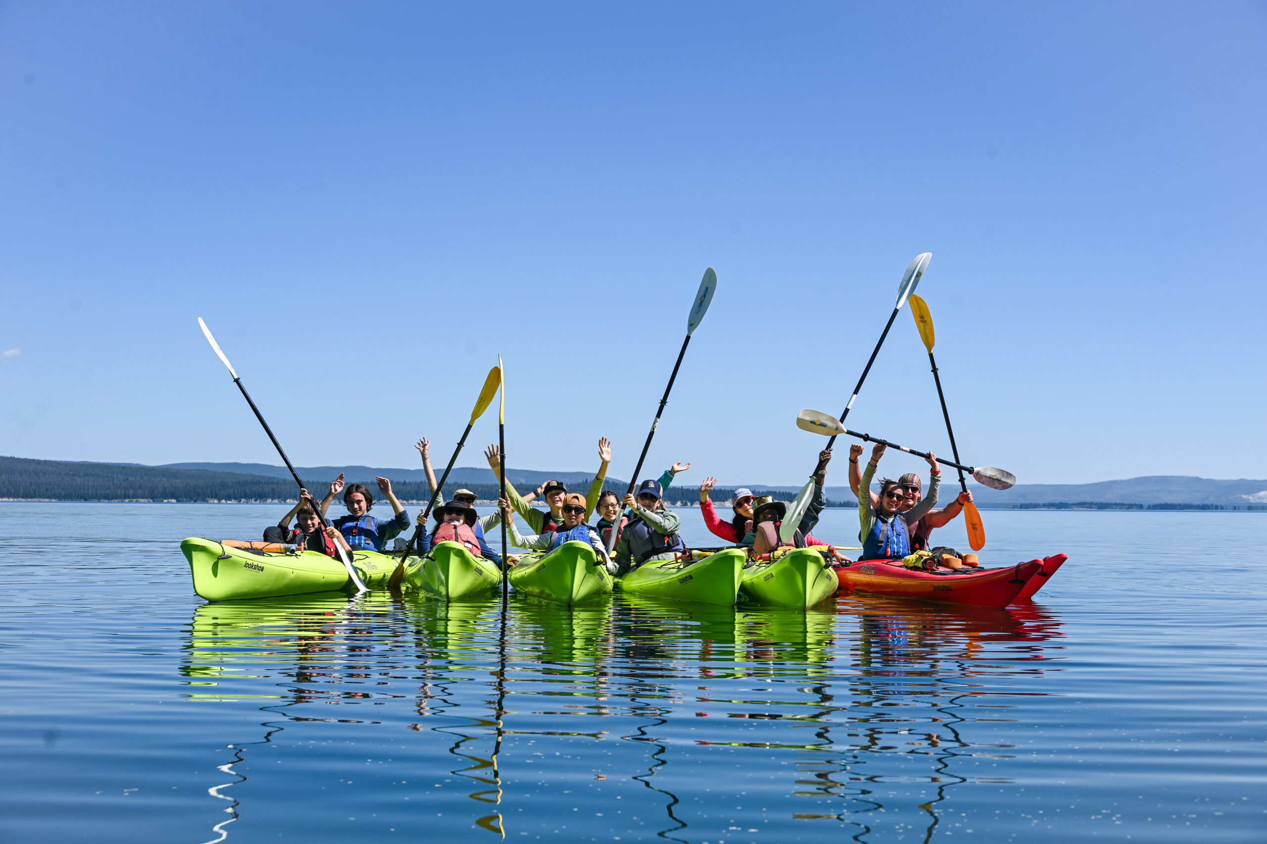 Group of students in kayaks on a blue lake holding their paddles in the air.