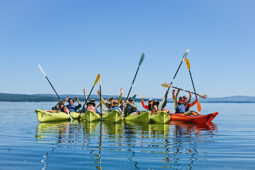 Group of students in kayaks on a blue lake holding their paddles in the air.
