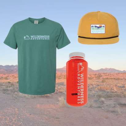 Wilderness Adventures Apparel - a green shirt, yellow hat, and red Nalgene water bottle all with the WA logo. Background is a desert landscape.