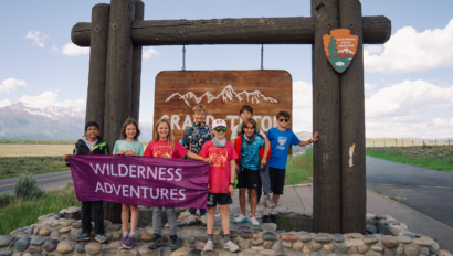 A group of summer camp kids in front of the Grand Teton National Park sign holding up a Wilderness Adventures banner.