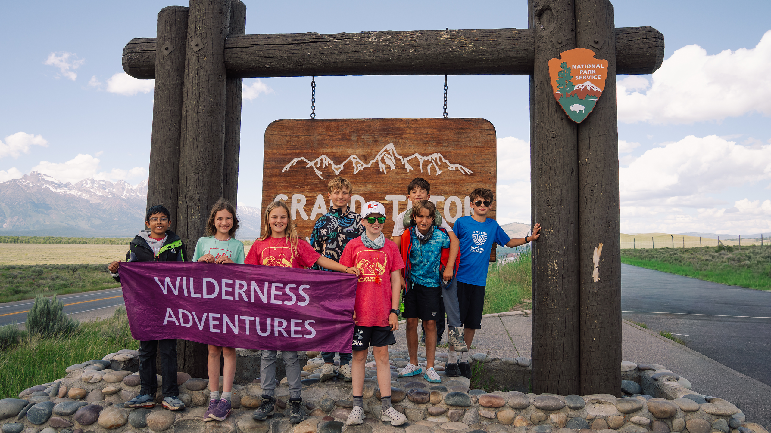 A group of summer camp kids in front of the Grand Teton National Park sign holding up a Wilderness Adventures banner.